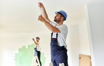 Young male electrician standing on ladders while installing spotlight during home renovation process.